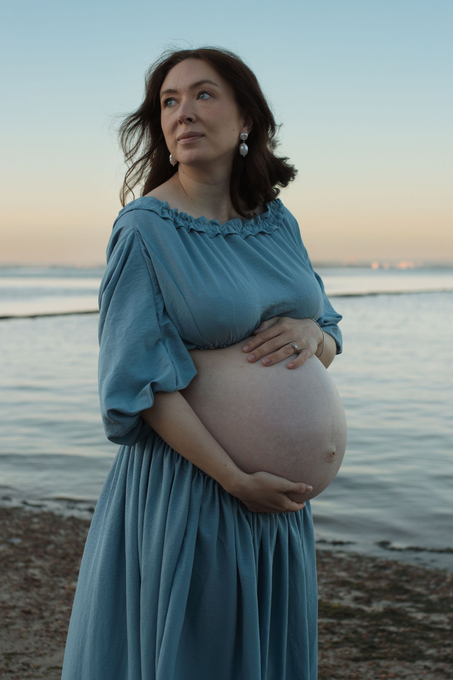 Pregnant woman in blue dress cradling her baby bump on a serene beach at dusk.