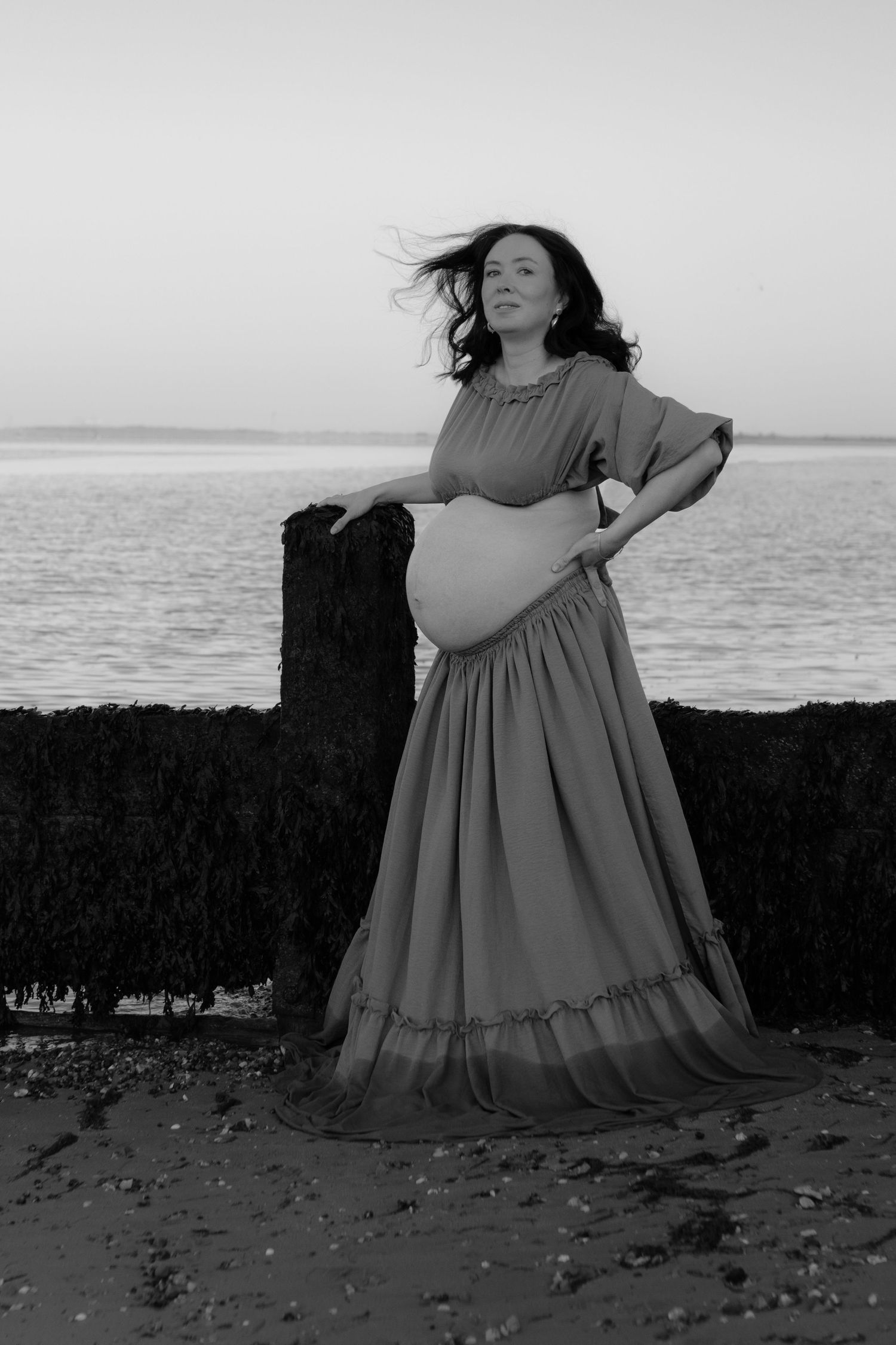 Black and white photo of a pregnant woman in a flowing dress standing on a beach, cradling her belly with the ocean behind her.