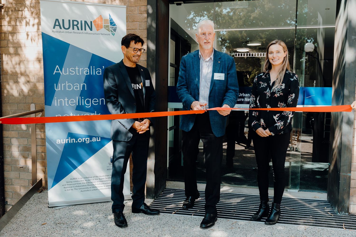 Three people cutting a ceremonial ribbon at a business opening event with an Aurin banner displayed.