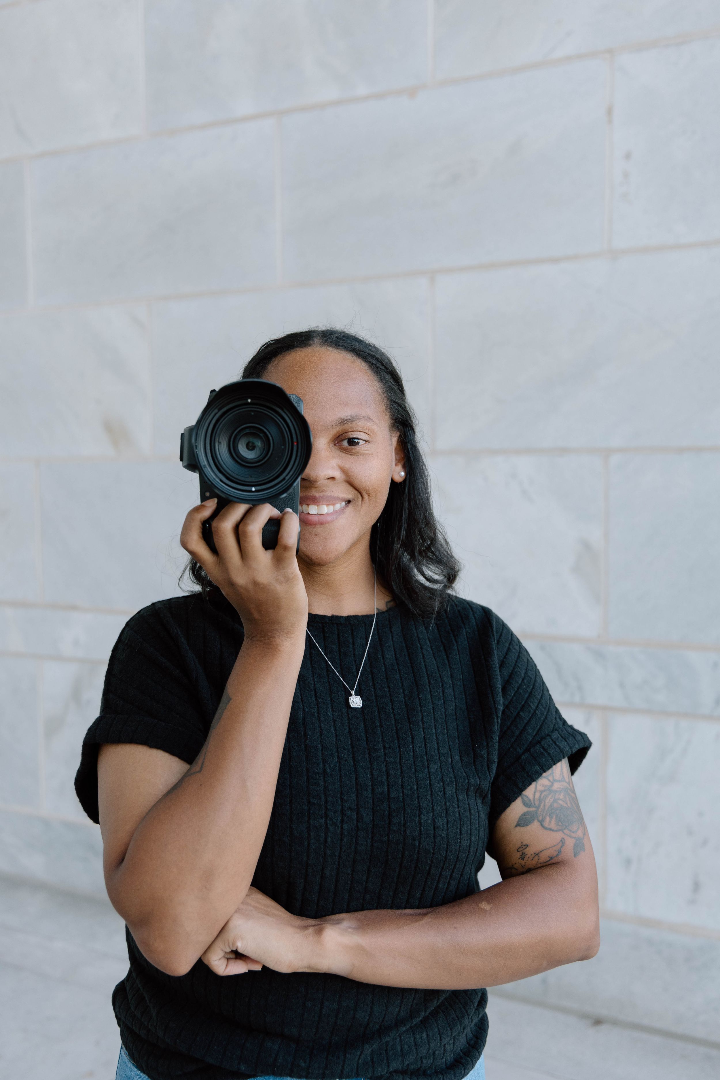 Professional photographer in black attire holding a camera against a white background.
