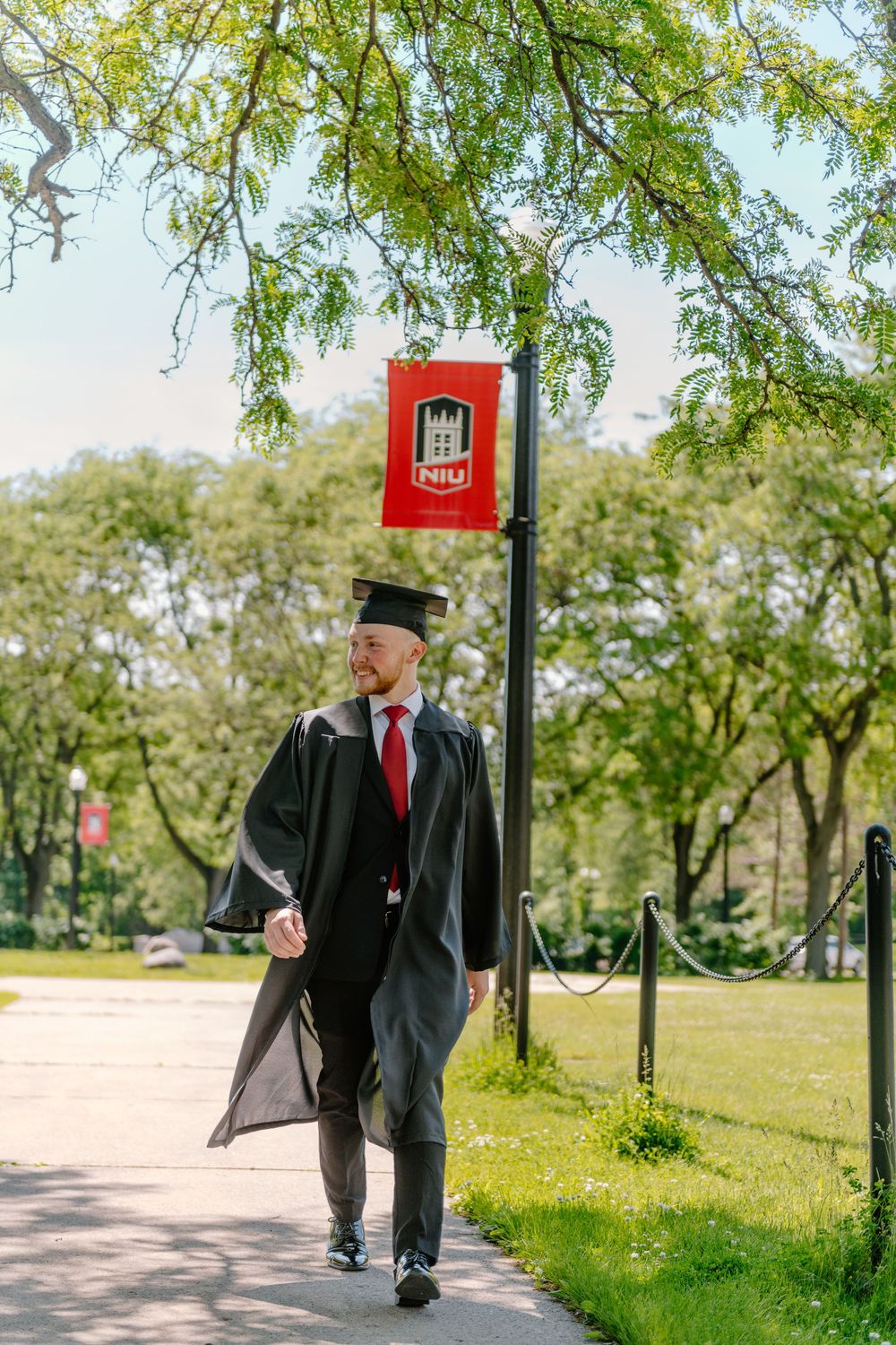 A graduate in a dark suit and red tie walks gracefully along a tree-lined path in a lush green park on a sunny day.