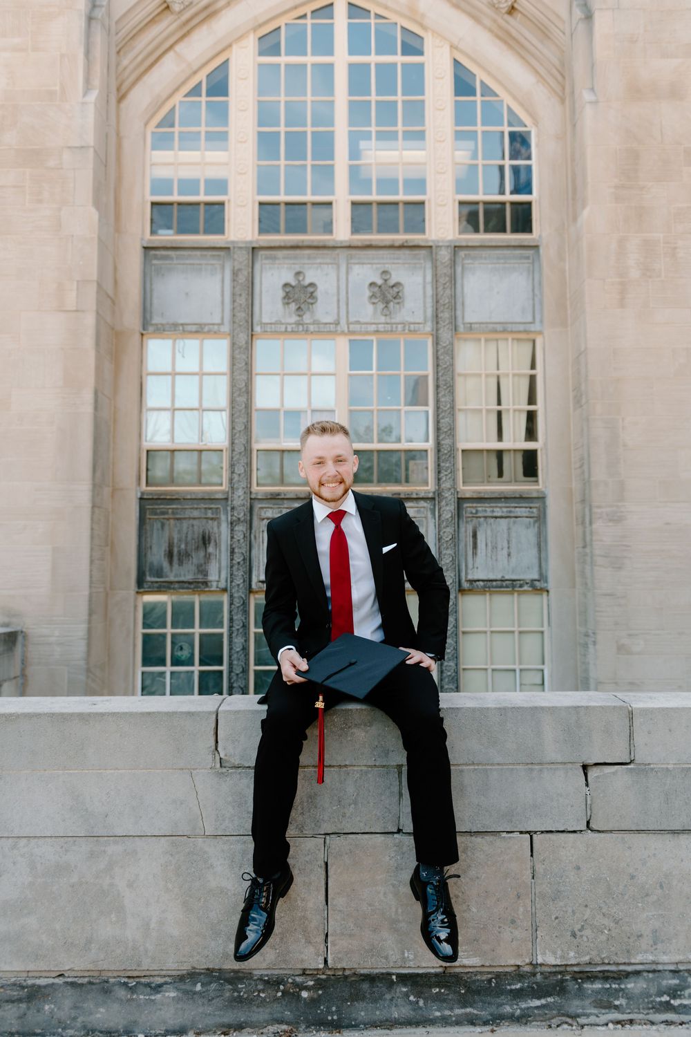 A graduate sits against a light stone wall with arched windows wearing a dark suit and red tie.