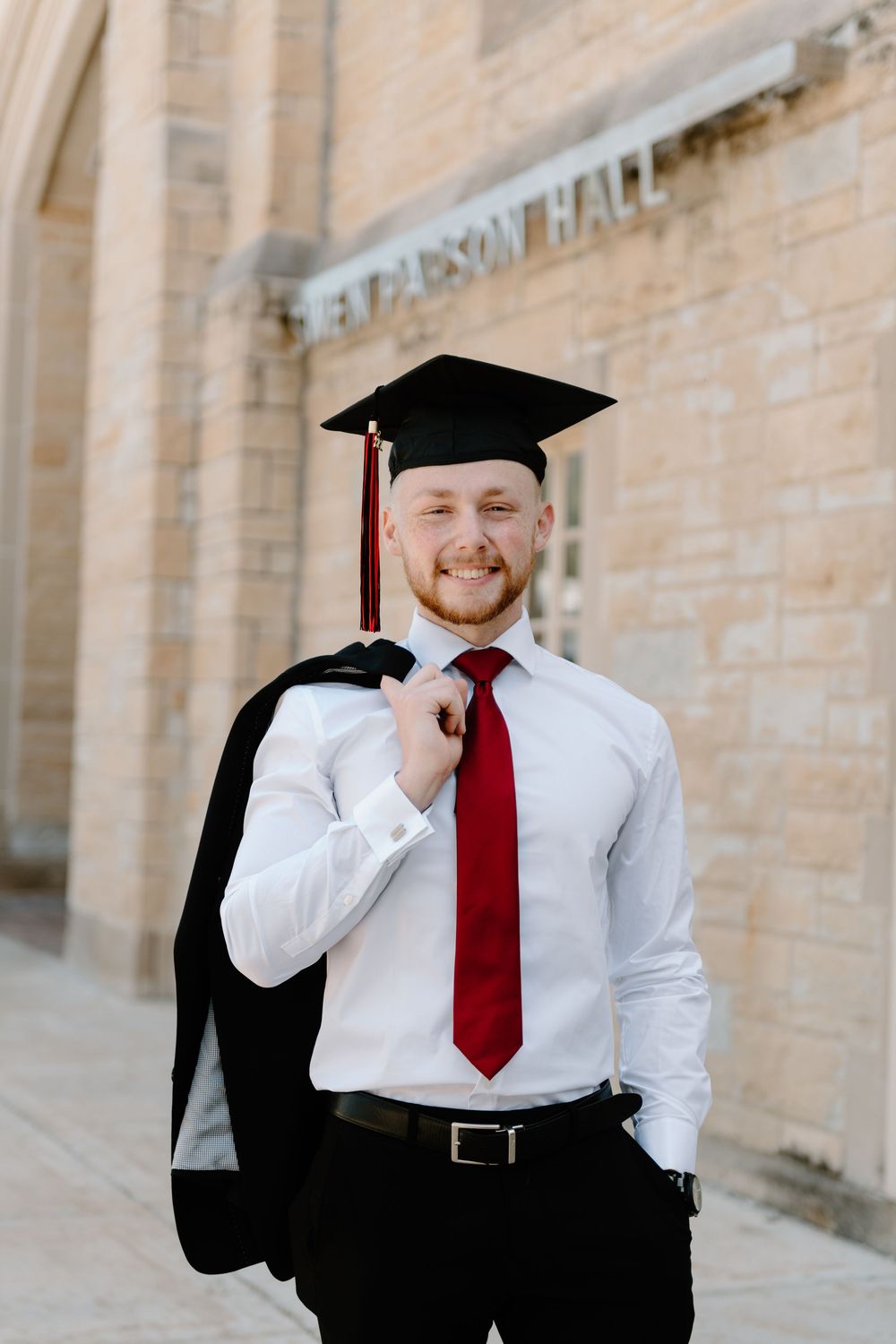 Graduate wearing a black cap and gown with red tie poses outside limestone building on graduation day.