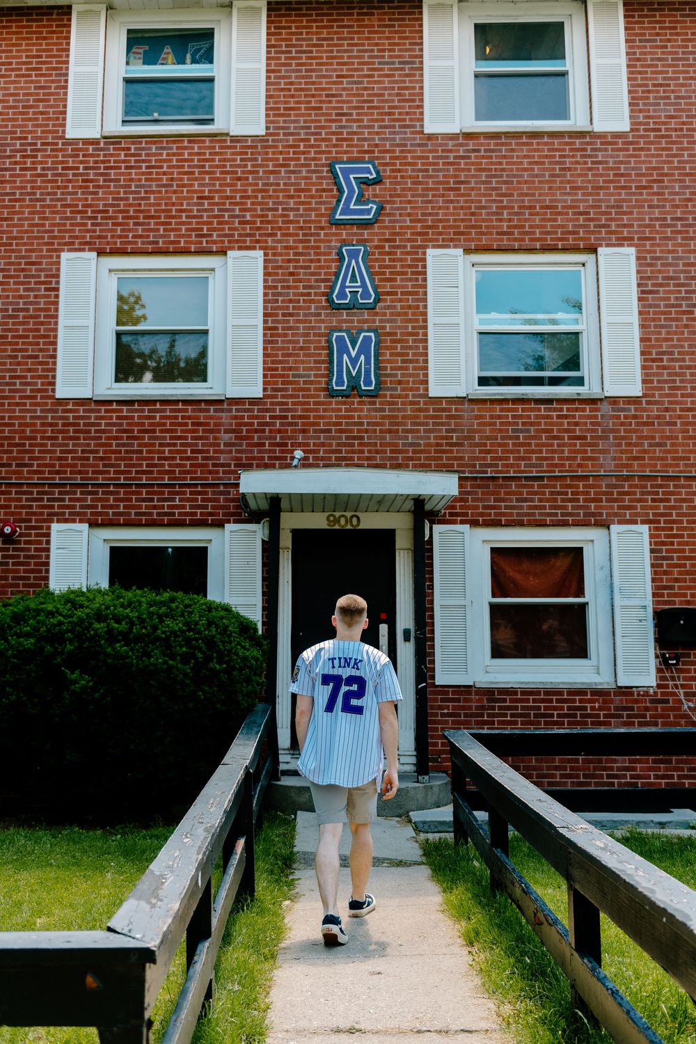 Someone in a sports jersey number 72 walking toward the entrance of a brick Sigma Delta Mu fraternity house.
