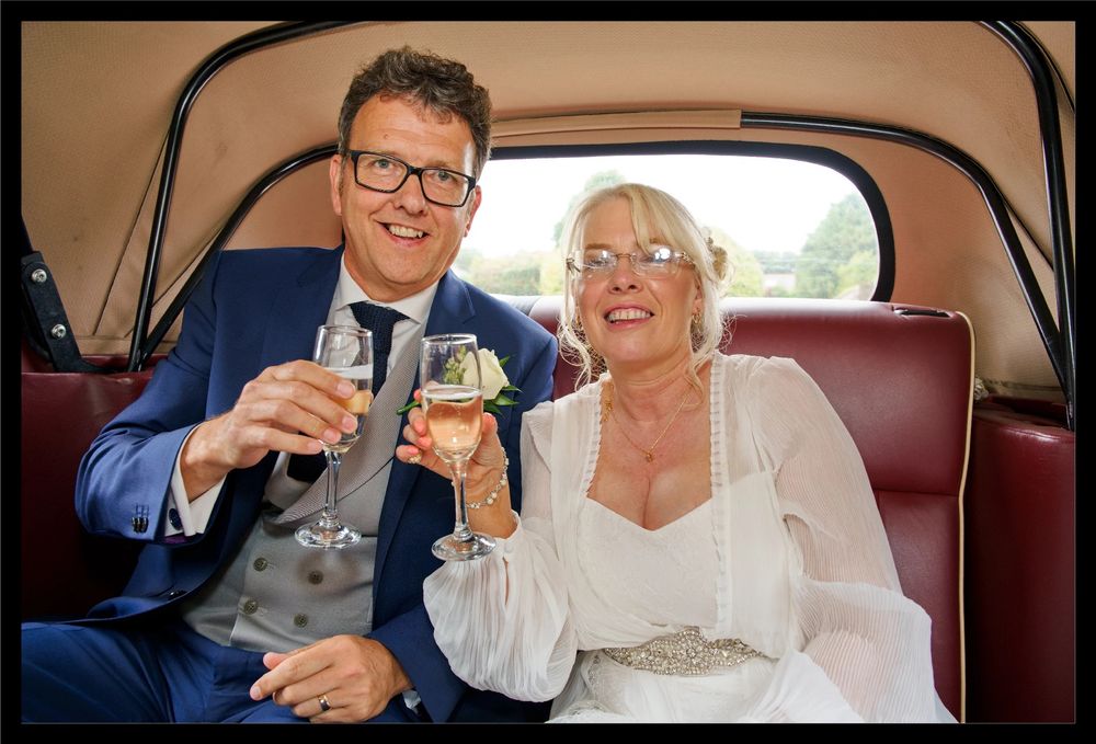 Bride and groom inside their wedding car, raising glasses in a celebratory moment.