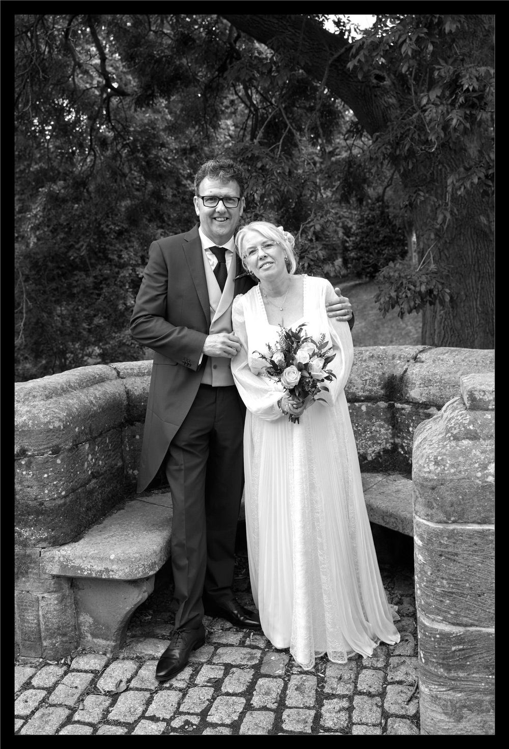 Black and white wedding photo showing a couple standing on stone steps beside an old wall with trees in background.