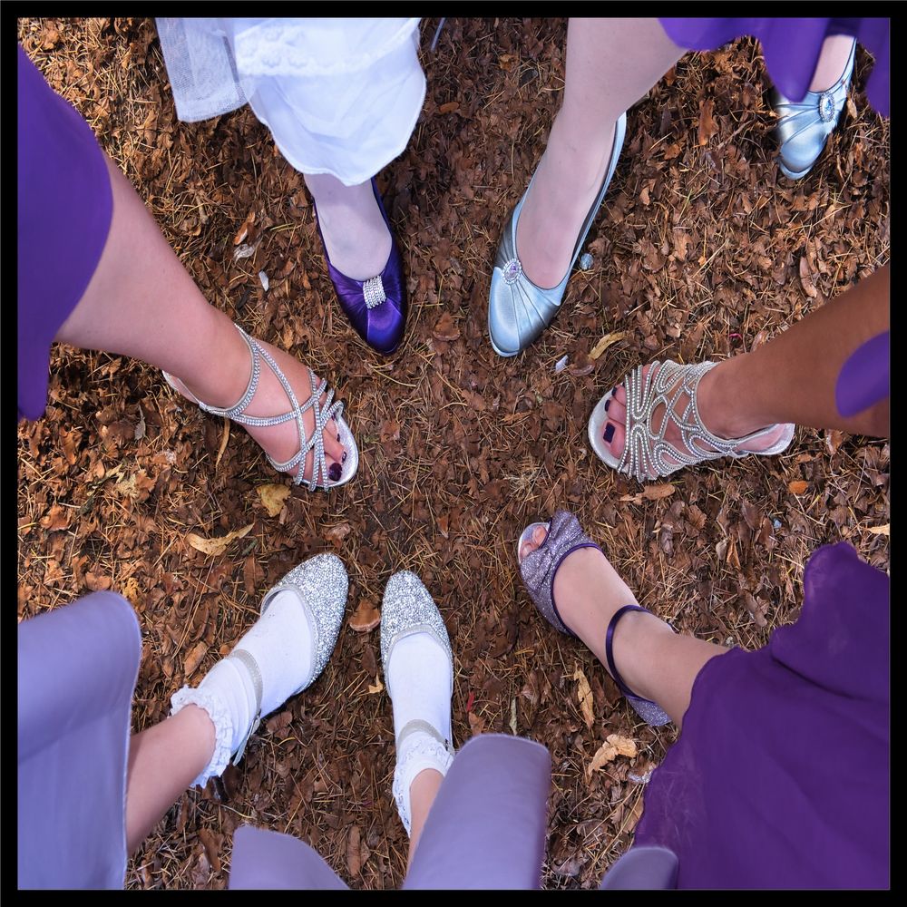Top-down view of six pairs of bride and bridesmaids' feet wearing different shoes and sandals arranged in a circle on brown mulch.