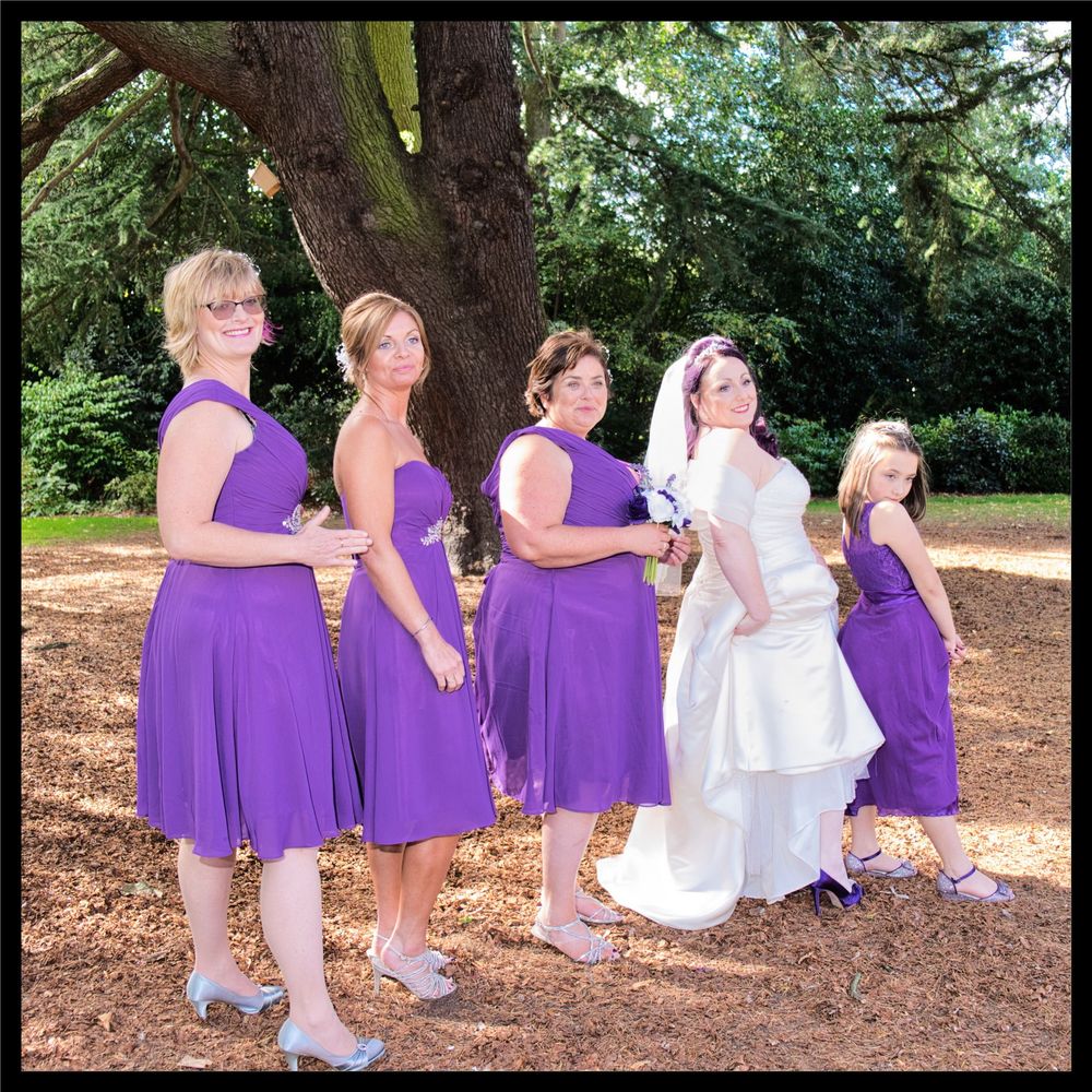 A bridal party in coordinating purple dresses poses in a line under a shady tree during an outdoor wedding.