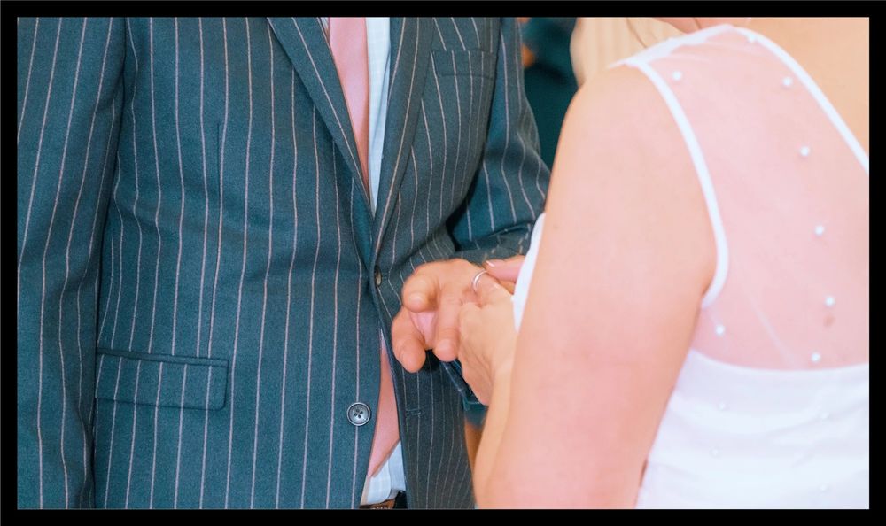 A cropped wedding photo showing hands holding together and formal attire in grey pinstripe and pink.