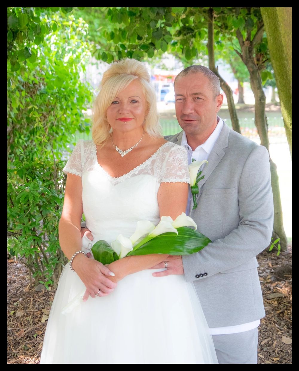 Elegant outdoor wedding photography shows a couple in white dress and light gray suit posing under green trees.