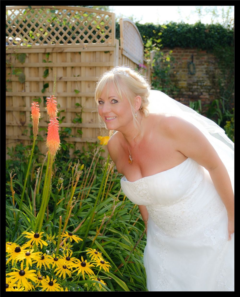A bride in a white wedding dress poses outdoors by a garden filled with yellow flowers and coral-colored plants.