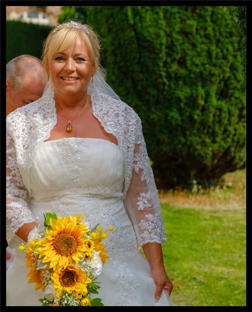 Smiling bride in white lace wedding dress holding vibrant sunflower bouquet poses outdoors on lawn with green hedge backdrop.