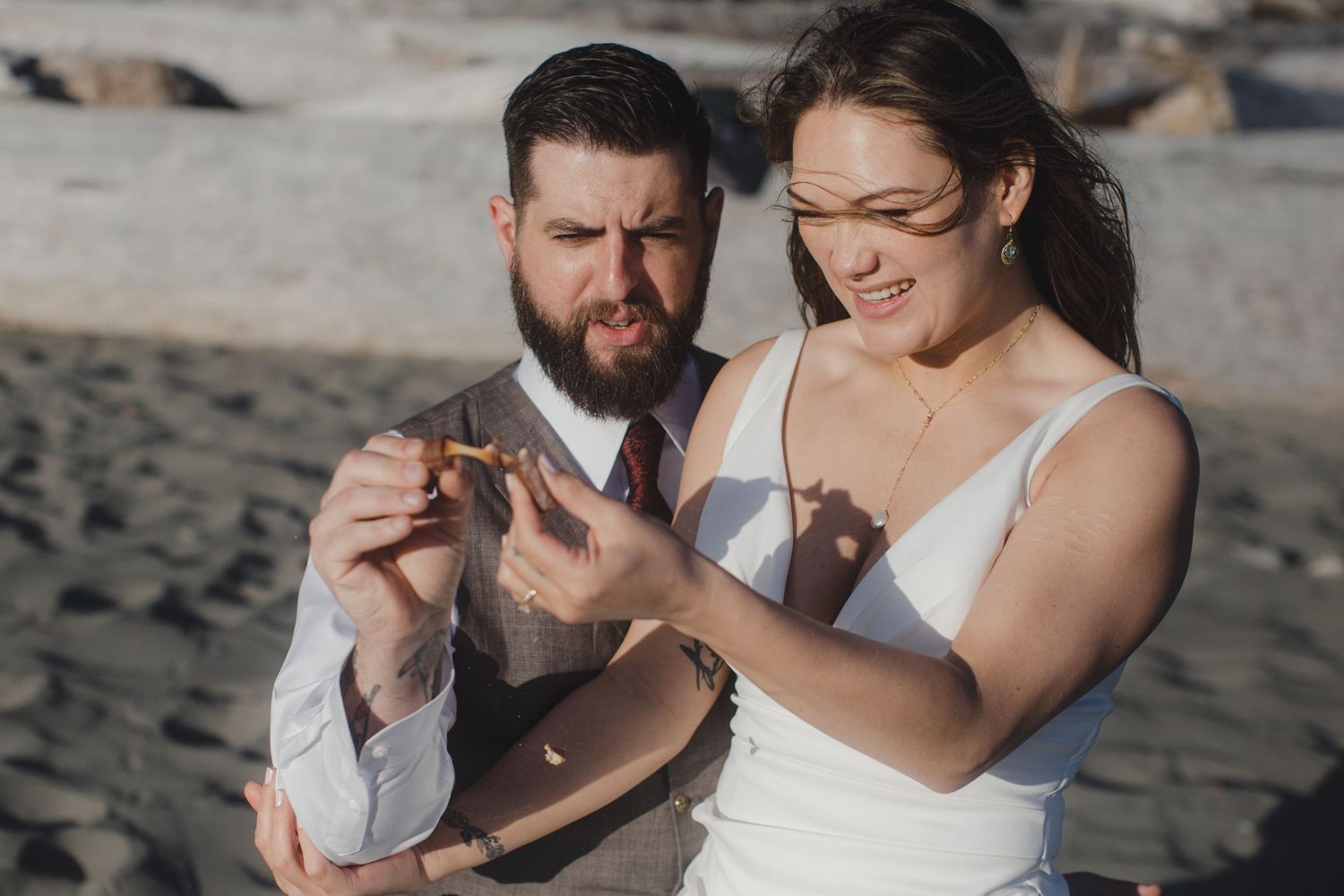 Couple sharing joyful moment while looking at something together on beach in casual wedding attire.
