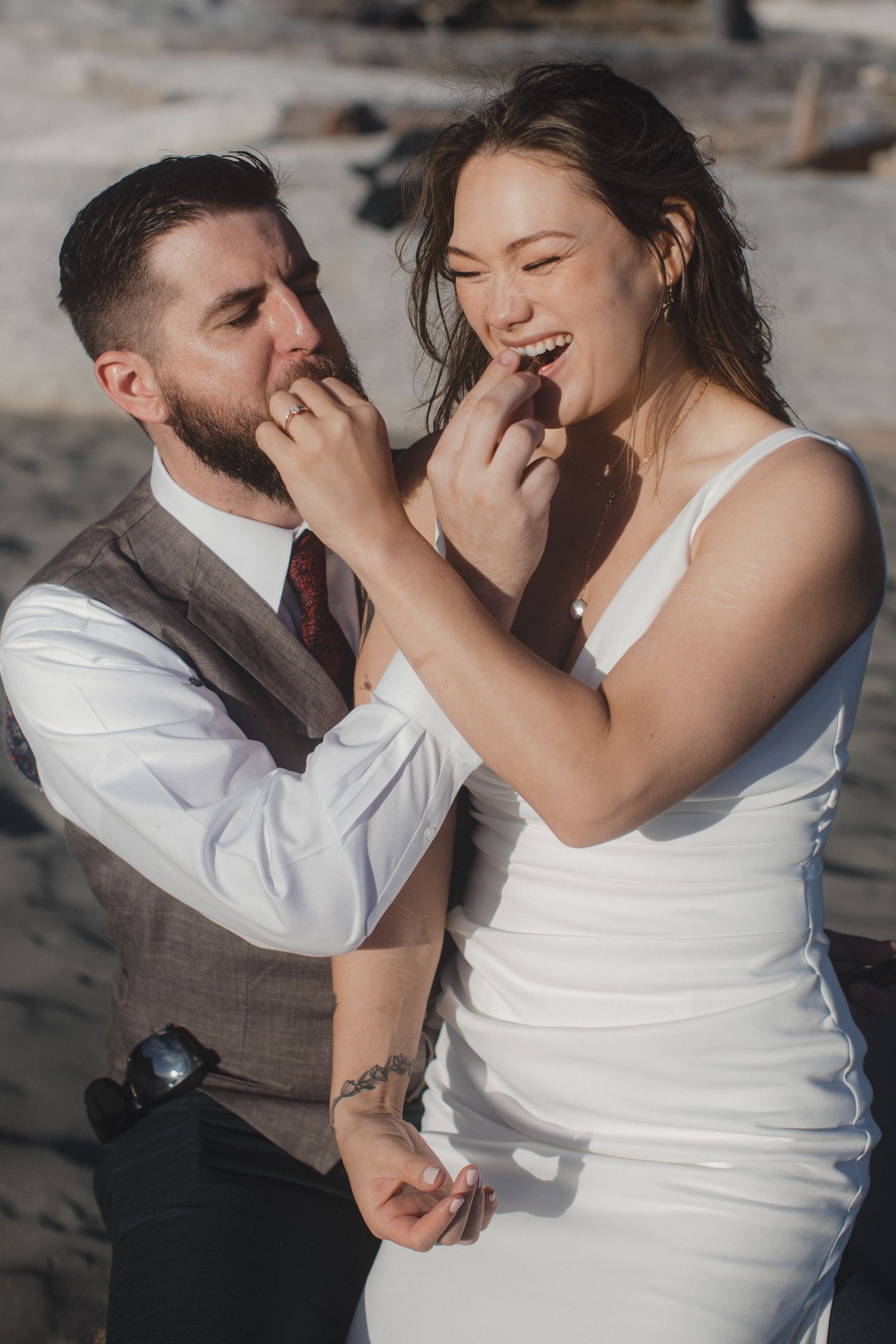 A couple in wedding attire laughs together while sharing a playful moment on a rocky beach.
