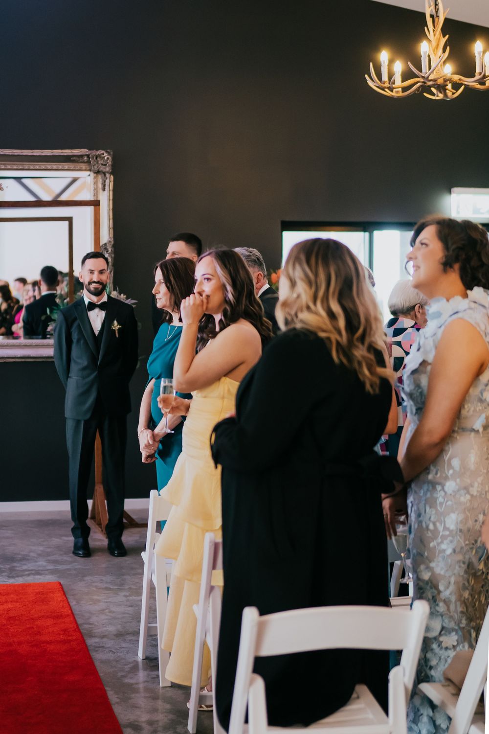 Guests at a formal event gather to socialize in a room with red carpet and elegant chandelier lighting.