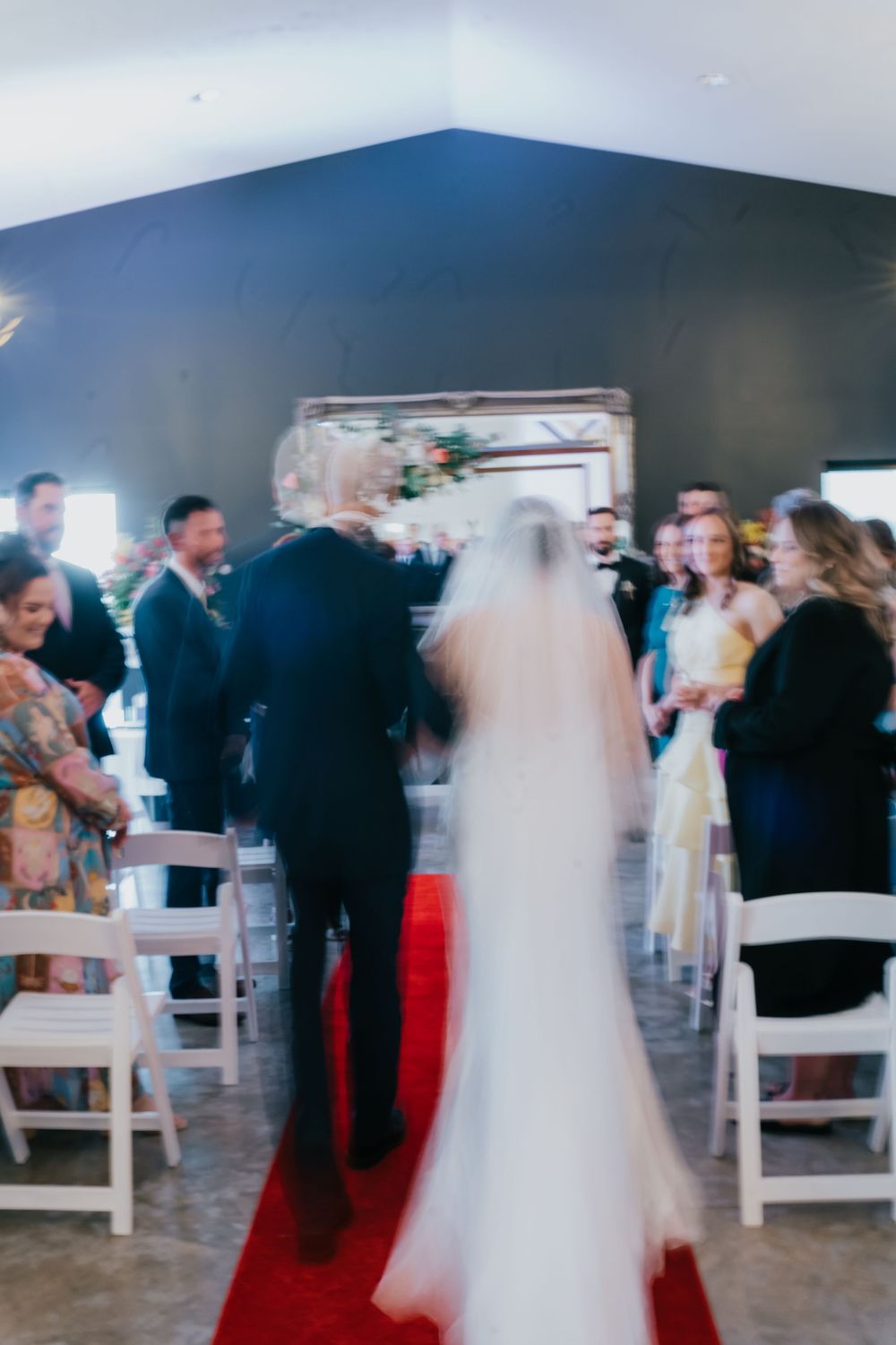 Blurred motion shot of wedding guests dancing and mingling in a reception venue with white chairs and dark walls.