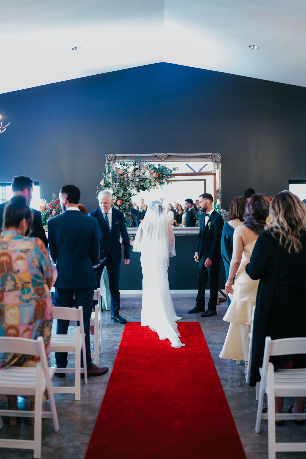 Wedding ceremony in modern space with red carpet aisle and white chairs as guests watch bride walk down the aisle.