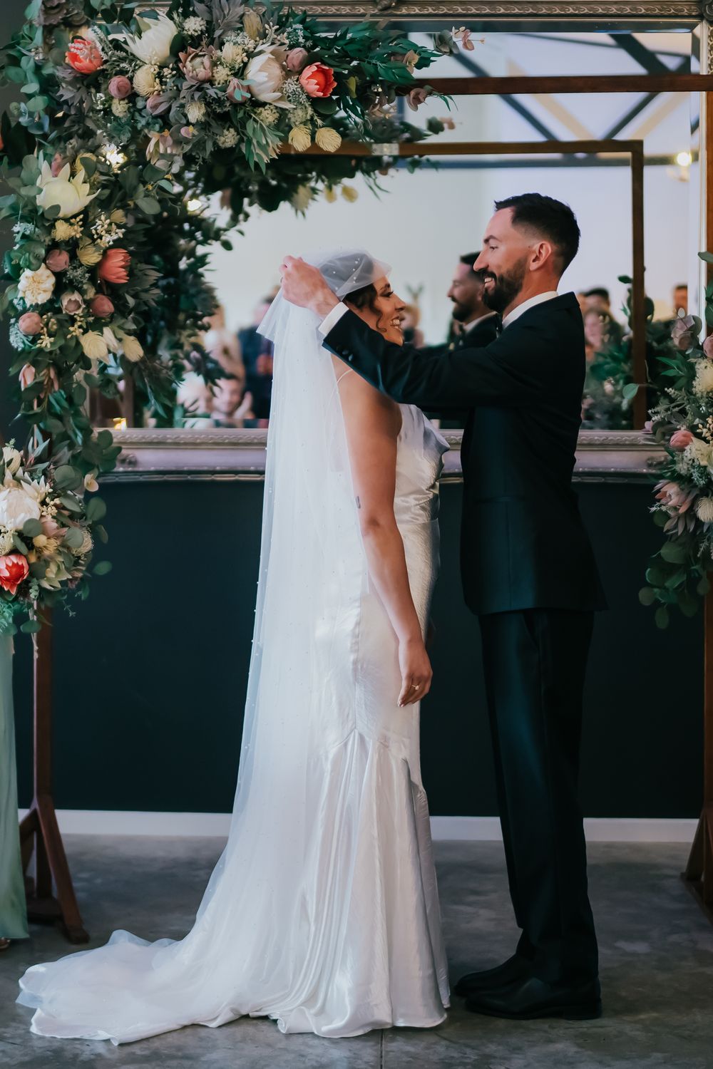 A couple shares a tender moment under a floral arch decorated with pink and white roses at their wedding ceremony.