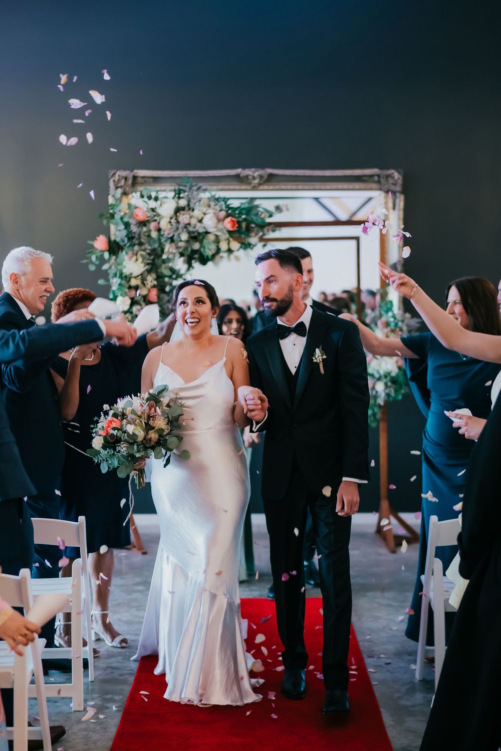 Joyful newlyweds celebrating during their wedding ceremony on a red carpet aisle with flower petals and white chairs.