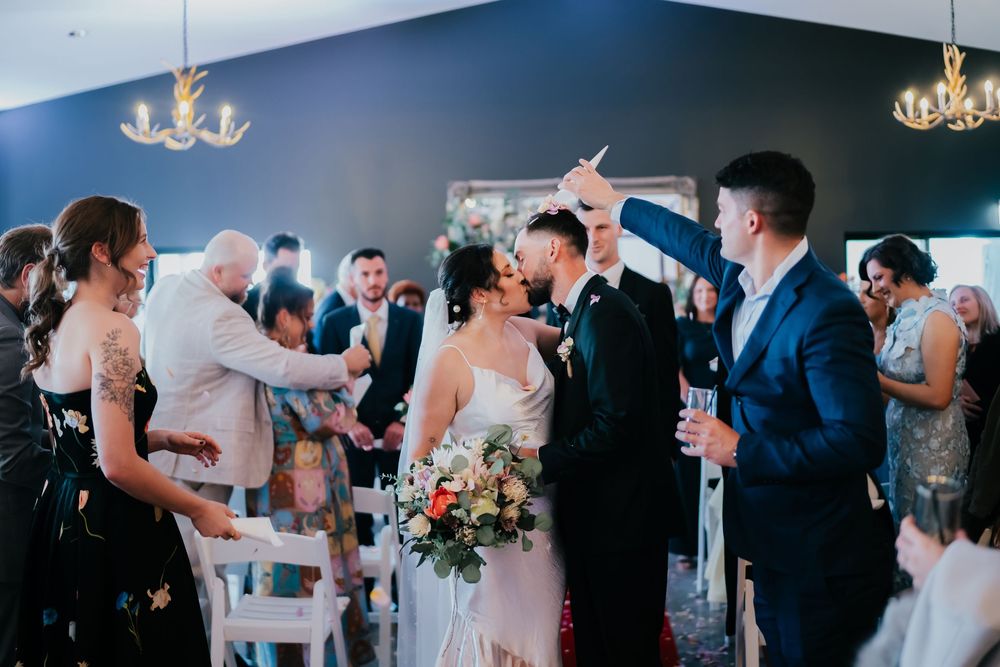 Wedding reception guests enjoying dancing in an elegant ballroom with chandeliers and dark blue walls.
