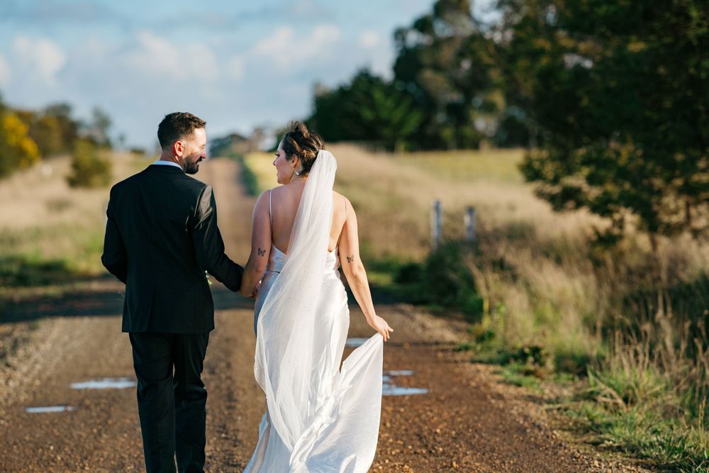A couple in wedding attire walking together on a dirt road surrounded by golden fields at sunset.
