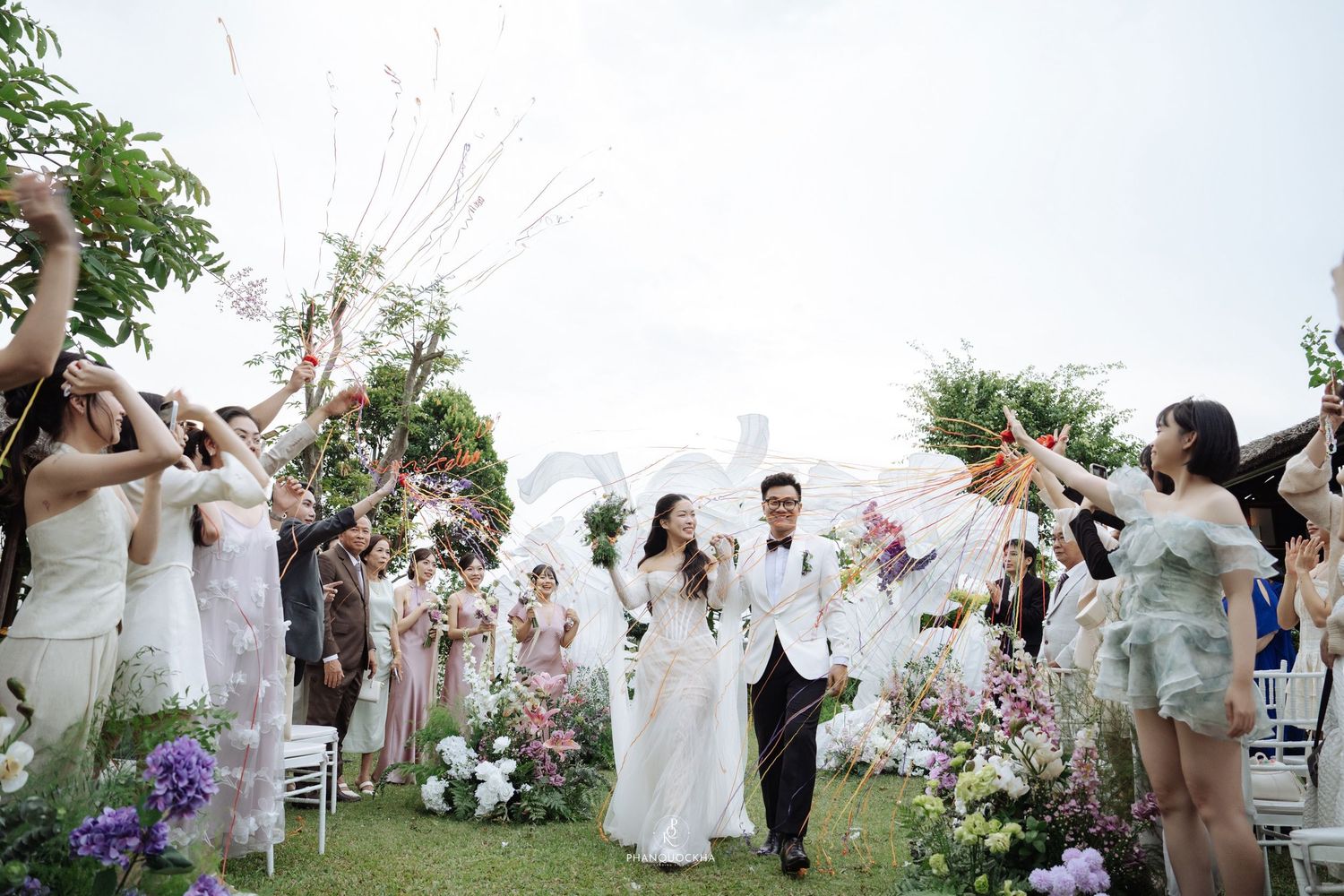 Newlyweds celebrate joyful outdoor wedding ceremony exit with guests cheering and holding flowers.