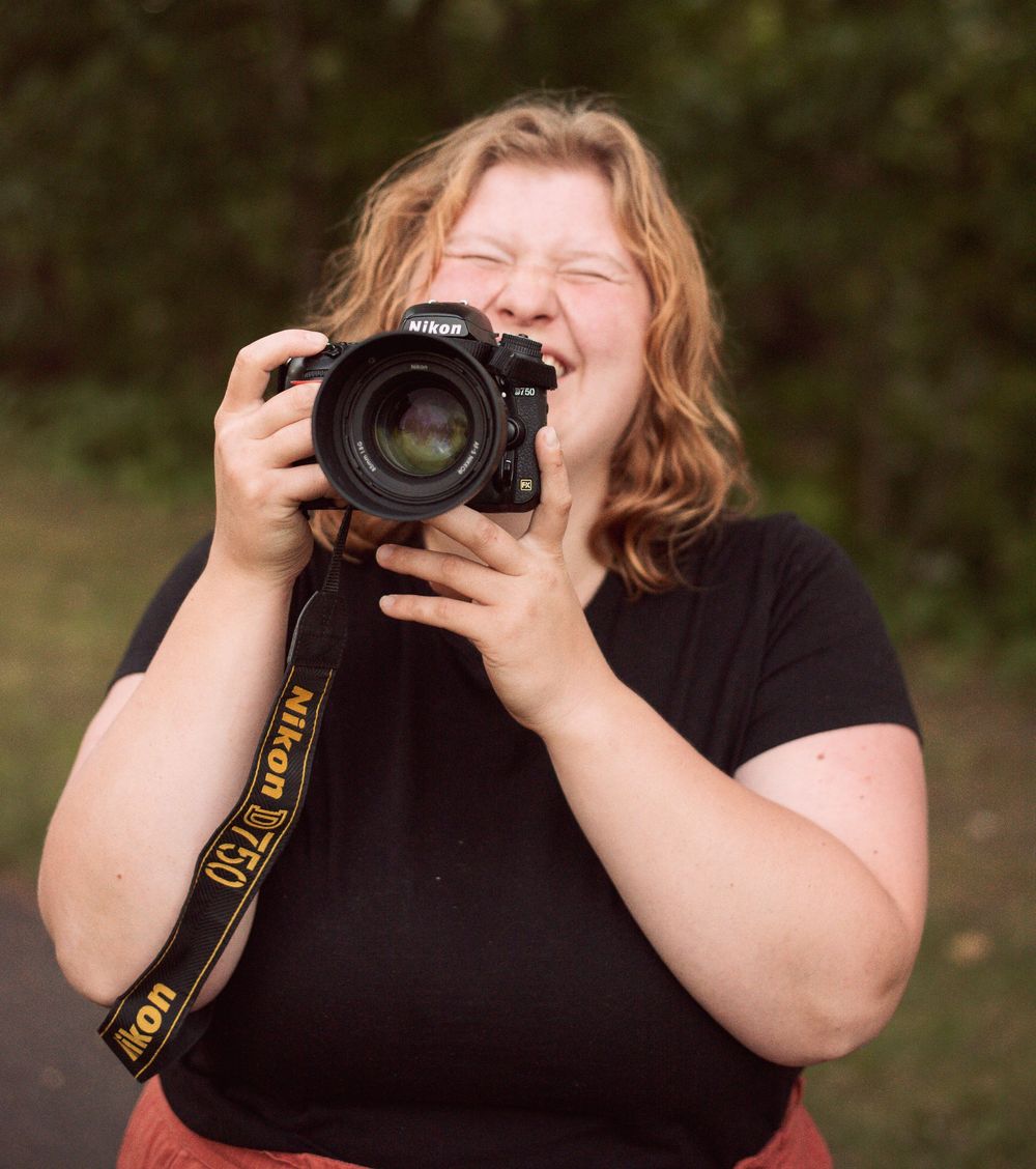 Laughing woman holding a Nikon D750 camera outdoors with green foliage in the background.