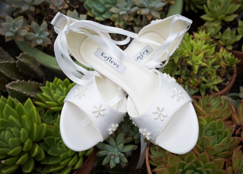 A pair of delicate white baby shoes rests among green succulent plants in a garden setting.