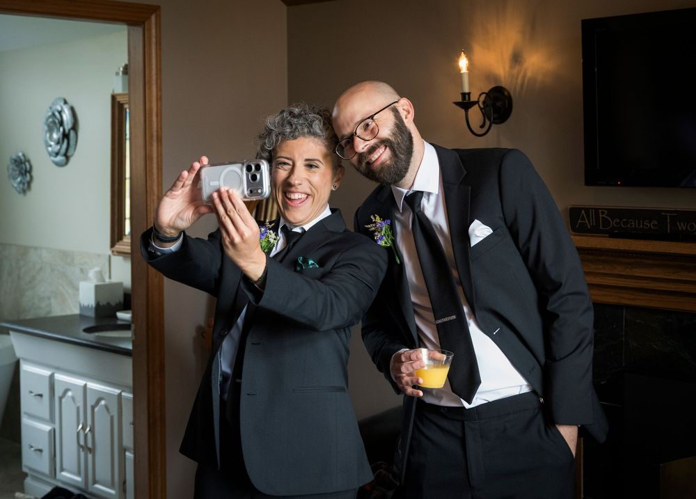 Two people in formal black suits pose together for a selfie at an elegant indoor venue.
