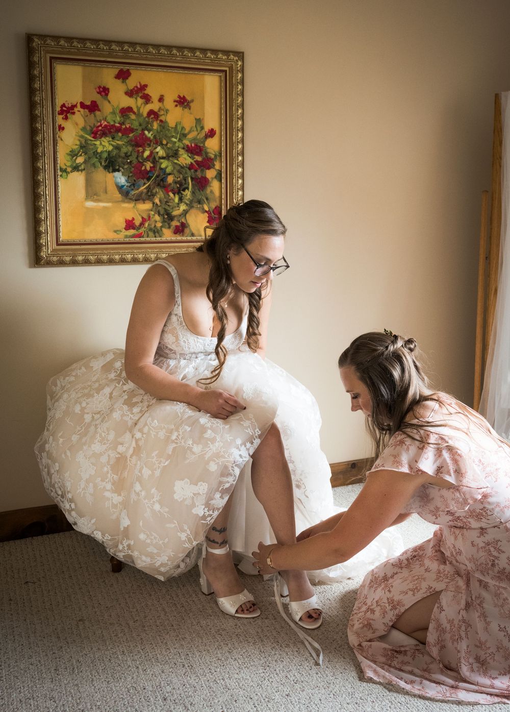 Bride getting ready with help adjusting her dress in a room with floral artwork on the wall.