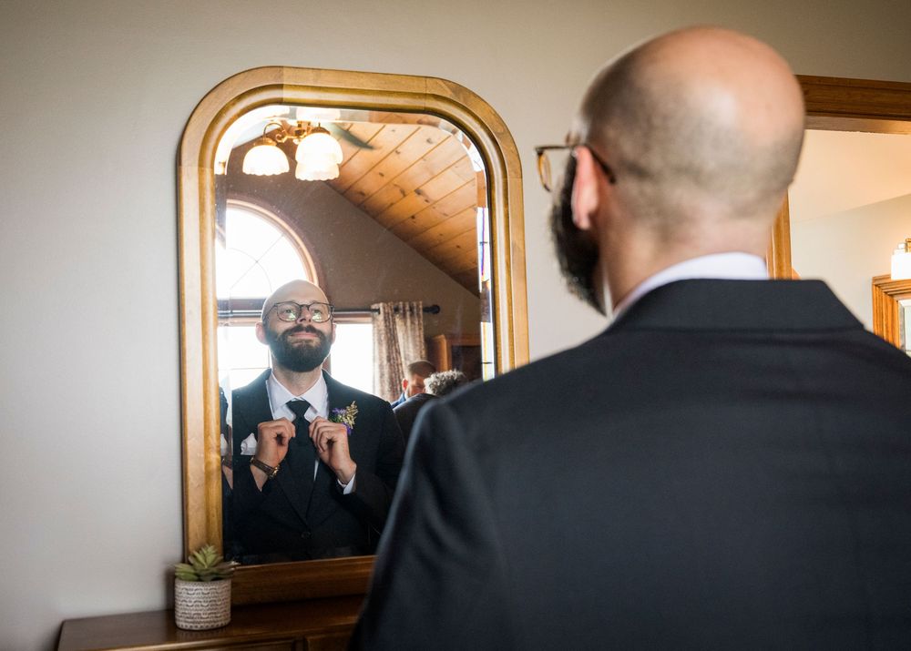 A person in a dark suit adjusts their tie while looking in a mirror in a warmly lit room.