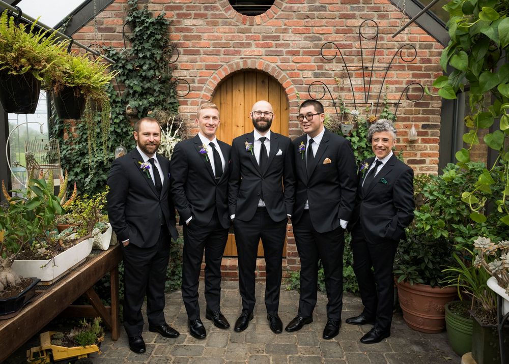 Wedding party in black suits posed against a rustic brick wall with arched doorway and ivy.
