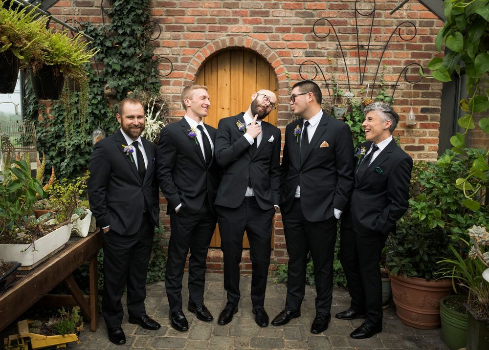 Wedding party in black suits posing against a rustic brick wall with wooden door.