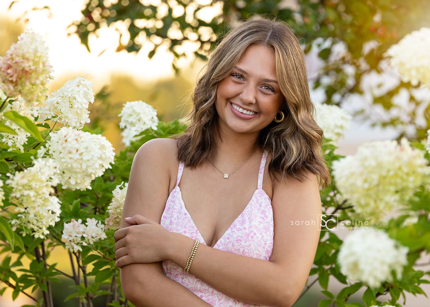 A golden sunset illuminates a figure in a pink top posing among blooming white hydrangea flowers in a garden.