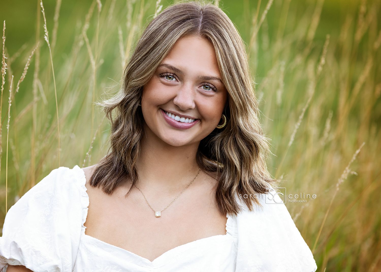 A smiling person in a white top poses outdoors in tall grass during golden hour.