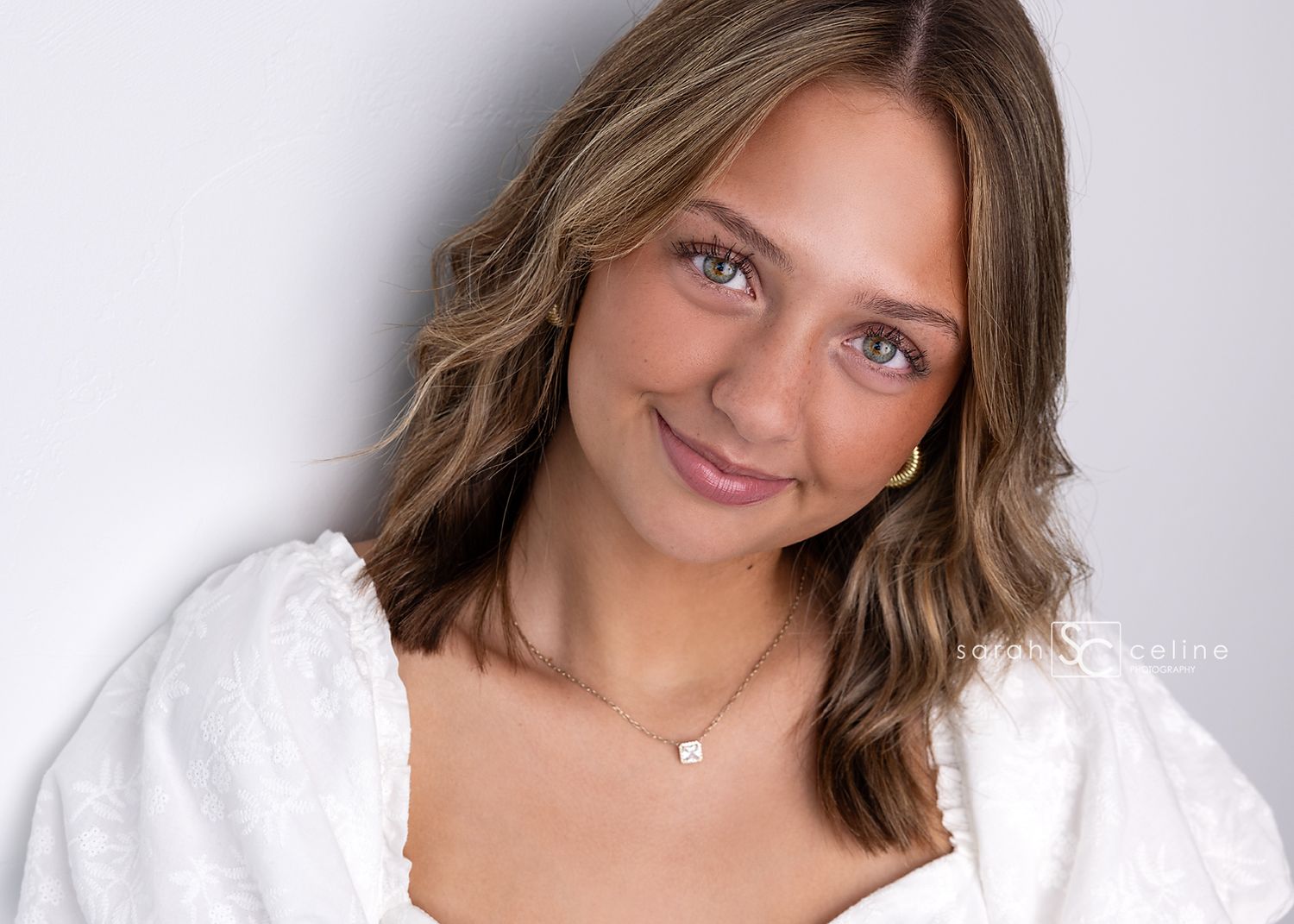 Portrait of someone wearing a white sweater and delicate necklace with natural lighting against a gray background.