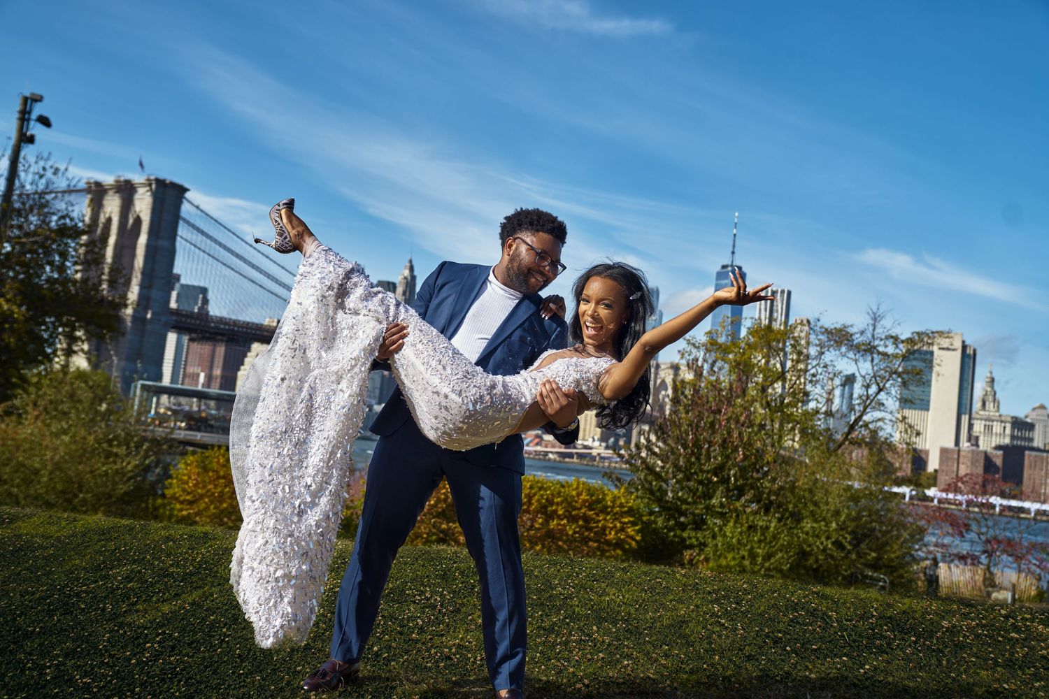 A joyful couple in wedding attire pose playfully at Brooklyn Bridge Park with the Manhattan skyline in the background at Brooklyn Bridge