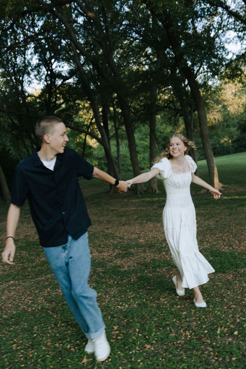 A couple in casual attire playfully dances together on fallen autumn leaves in a park setting.