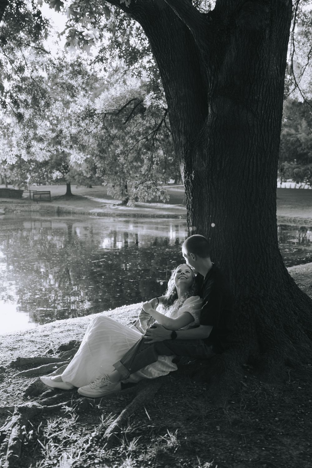A couple relaxes under a large tree in a serene park setting with a lake in the background.