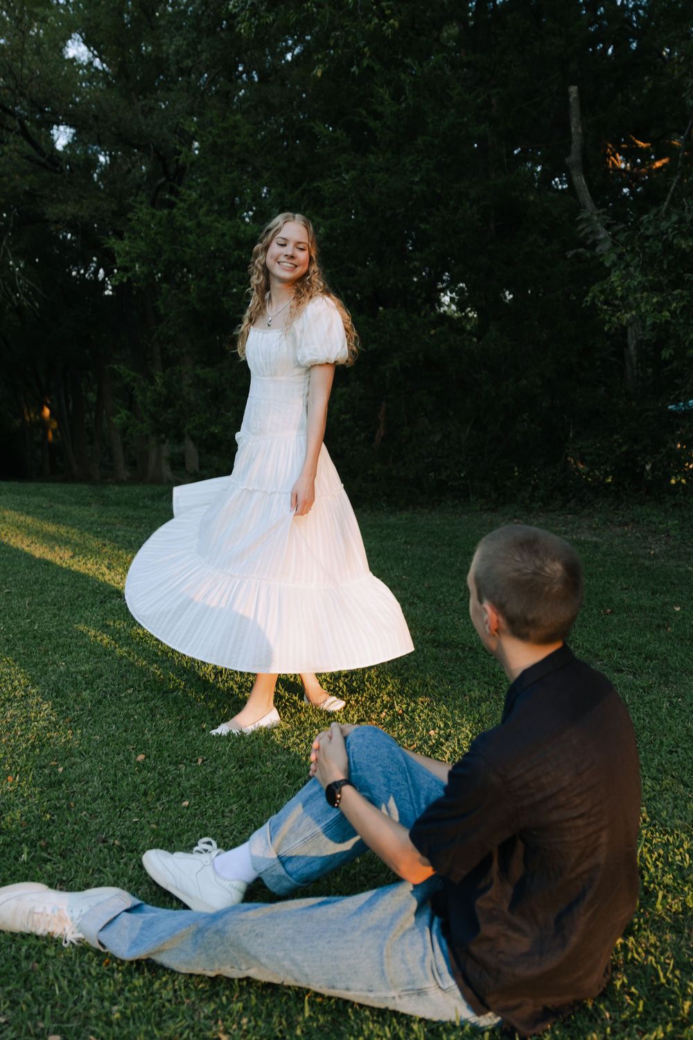 A couple shares a playful moment on grass during golden hour as sunlight filters through trees.