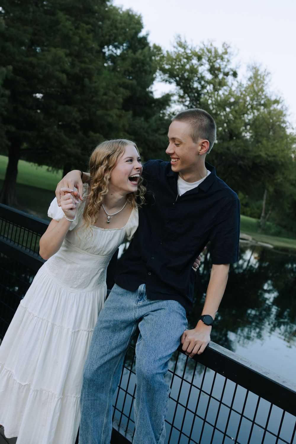 Couple laughing together while sitting on metal railing near a serene pond.