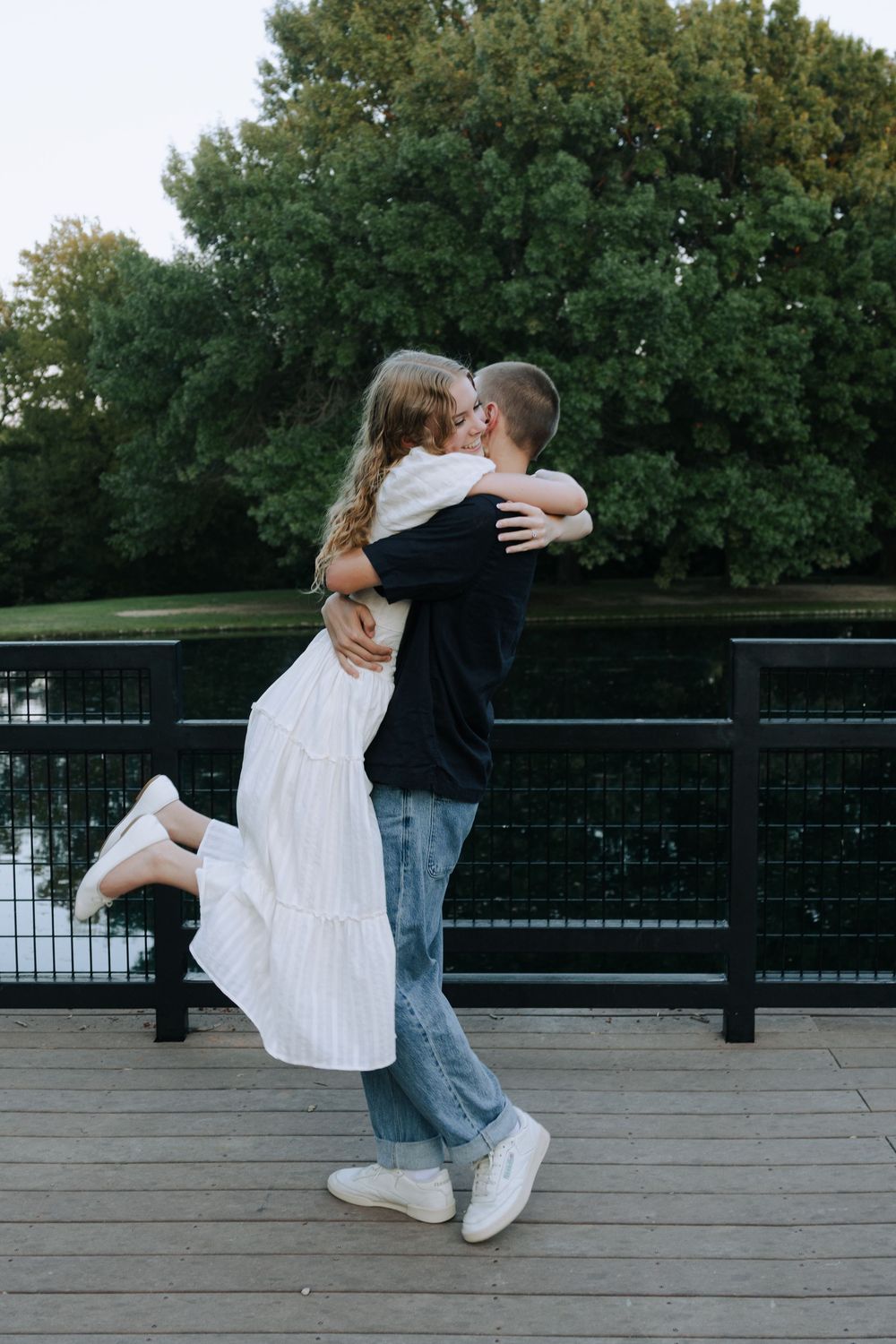 A couple in white dress and dark clothing embrace and dance together on a wooden deck surrounded by trees.