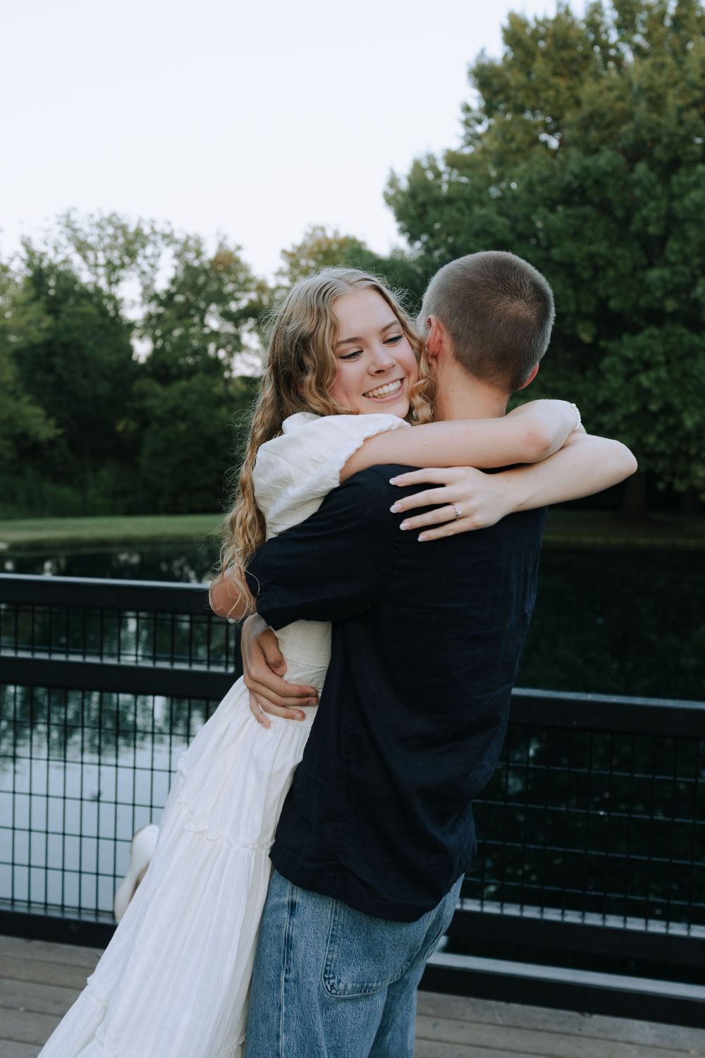 A couple shares a tender embrace on a bridge with trees in the background during a romantic outdoor photoshoot.
