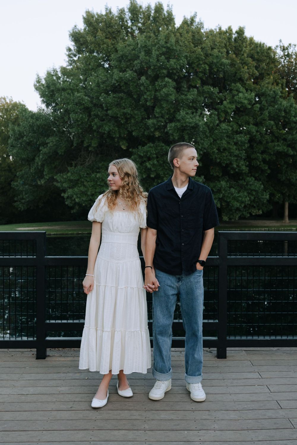 A couple stands together holding hands on an outdoor deck with trees in the background.