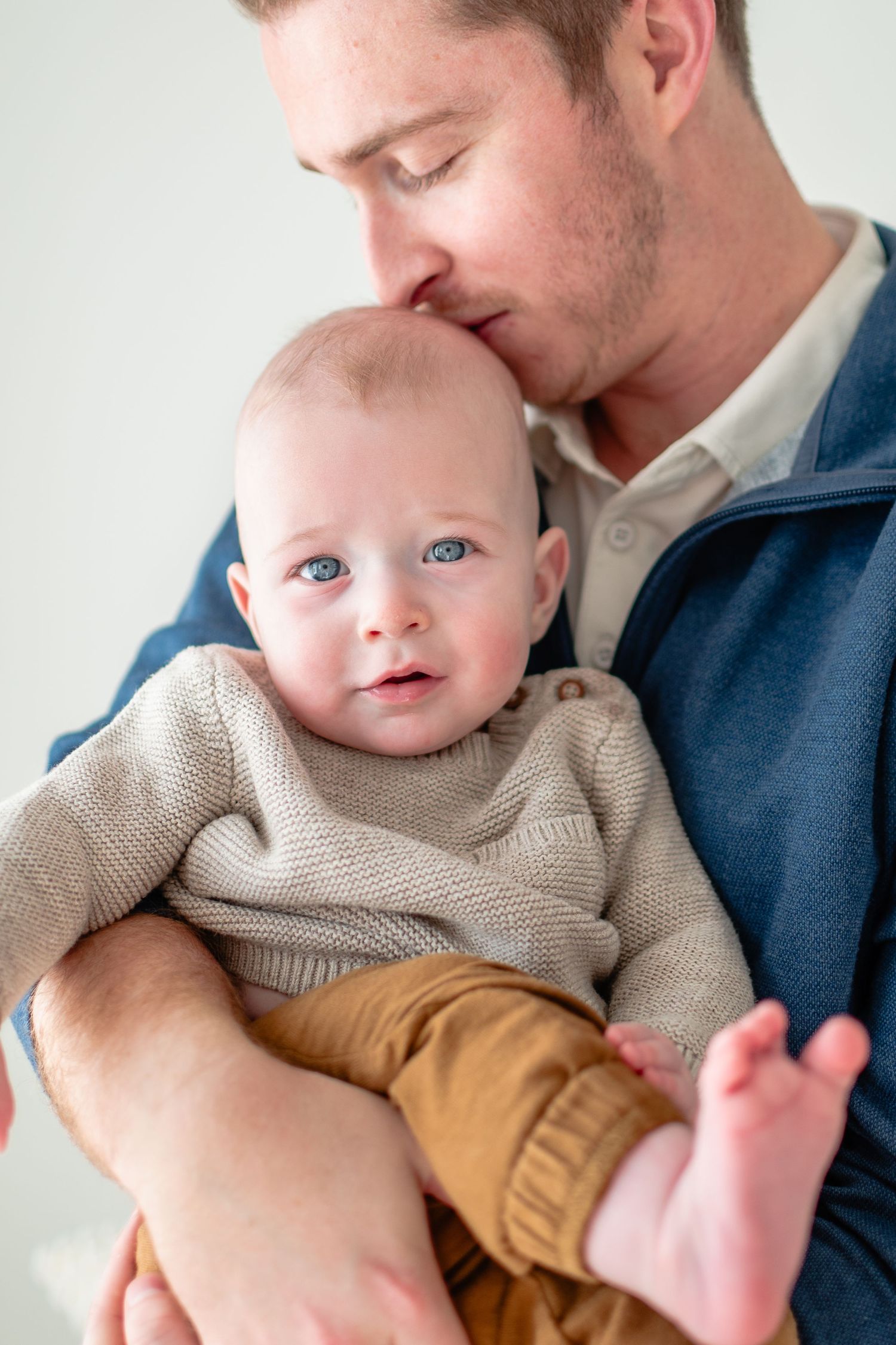 Baby in gray sweater and yellow pants smiles while being held against blue shirt background.