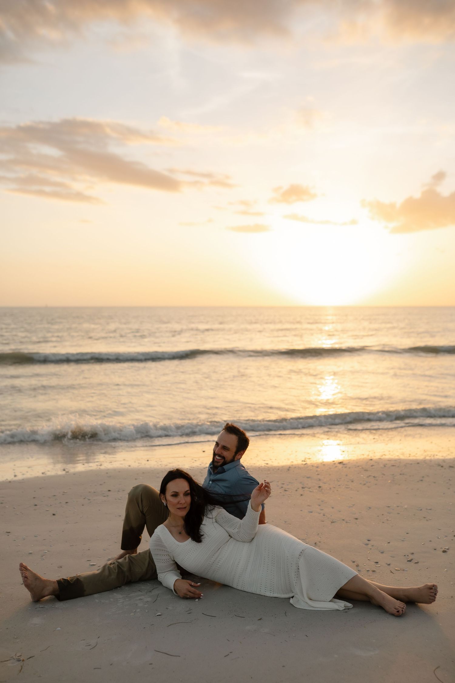A couple relaxes on a sandy beach at sunset with gentle ocean waves and warm golden sky in the background.