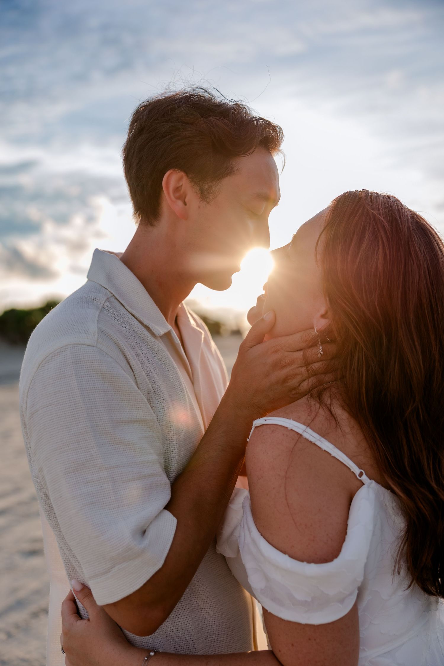 Romantic couple embraces on beach at sunset with sun creating golden backlight through clouds.