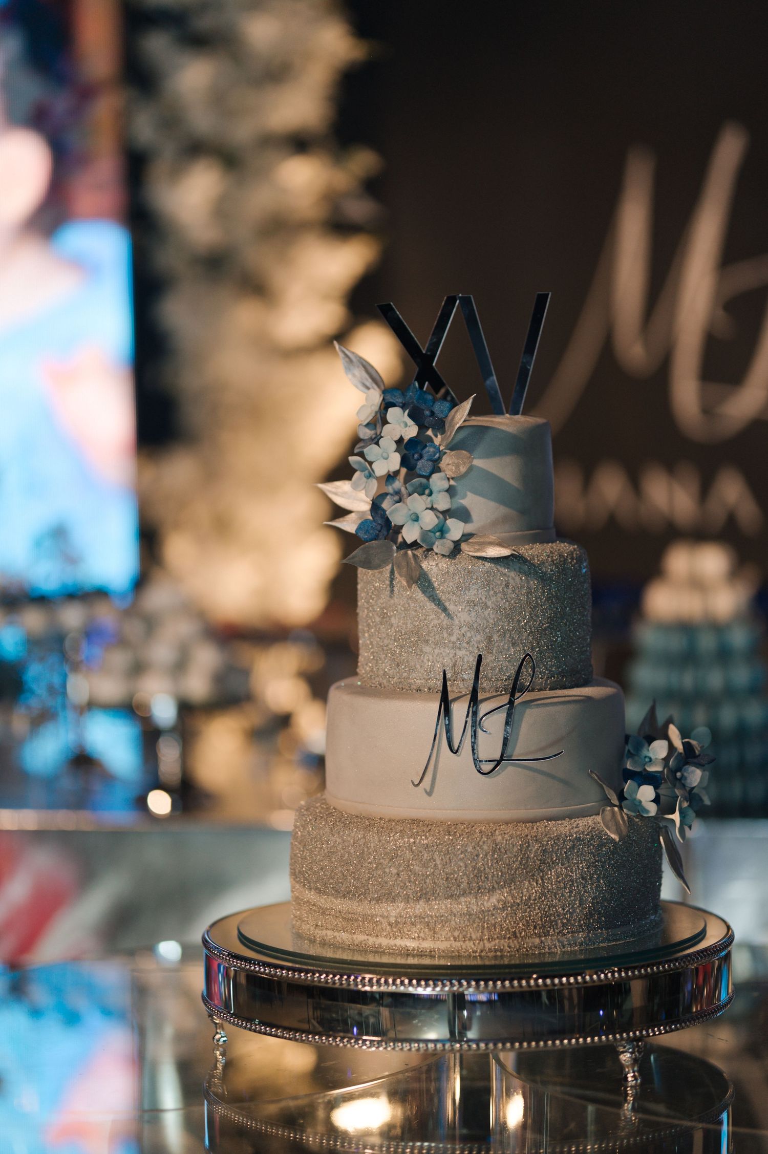 Elegant three-tier wedding cake decorated with silver glitter, monogram initials, and blue floral accents on a mirrored stand.