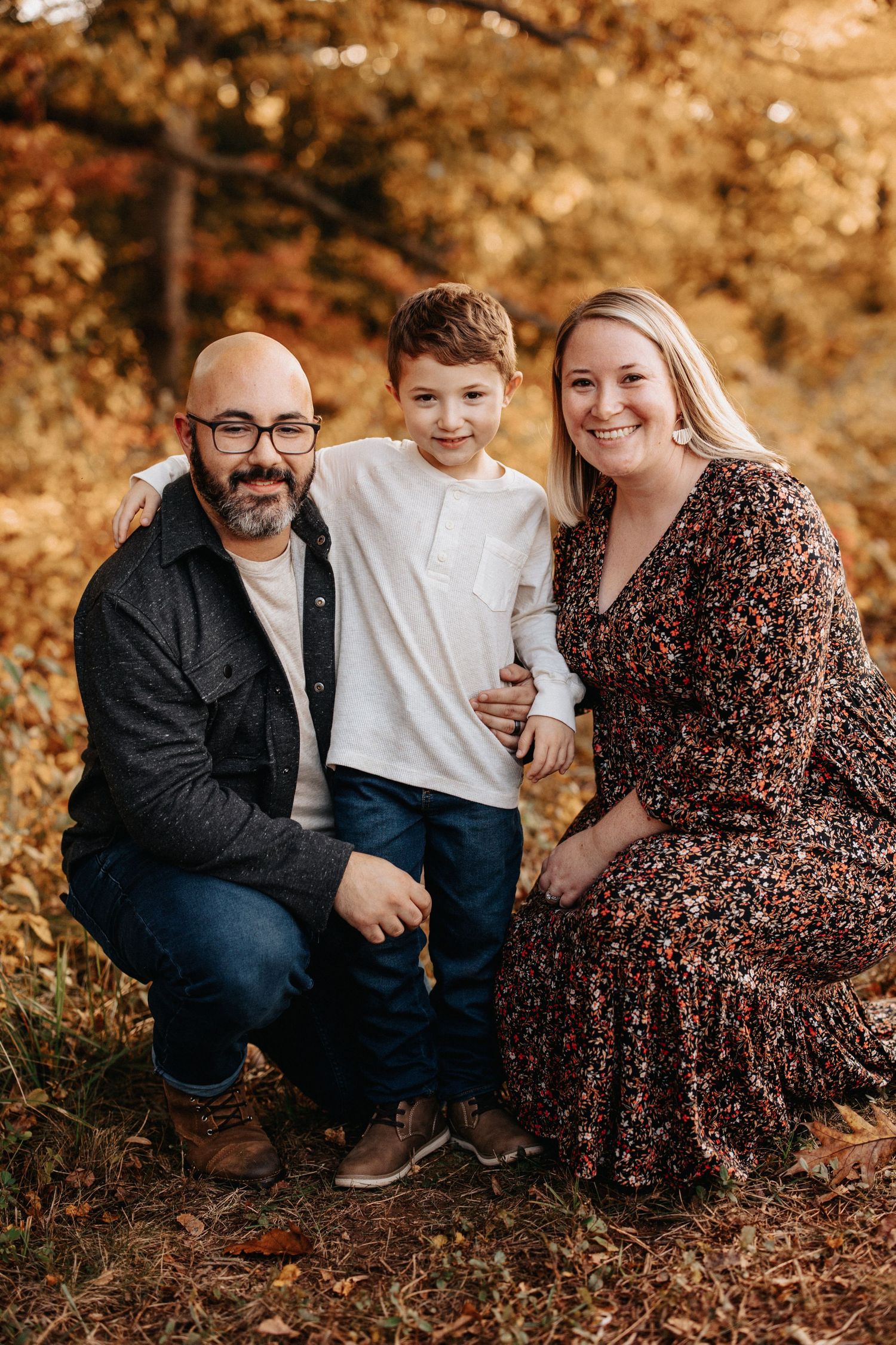 A family poses together outdoors in autumn with warm golden lighting and fall foliage in the background.