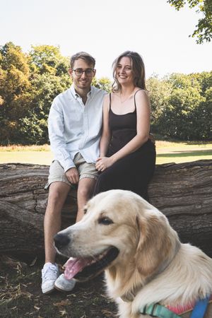 A couple sits on a fallen log in a park with their smiling Golden Retriever dog on a sunny autumn day.